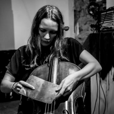 Leila Bordreuil, a thirty something white woman, playing the cello with a large piece of scrap metal placed under the strings on her fingerboard.